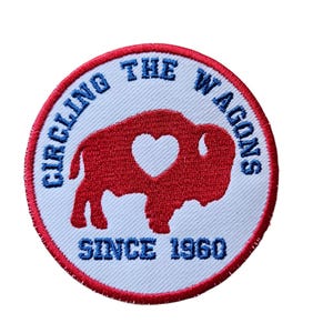 May include: A white, round embroidered patch with red and blue stitching. The patch features a red buffalo silhouette with a white heart inside. The text "Circling the Wagons Since 1960" is written in a blue circular font around the buffalo.