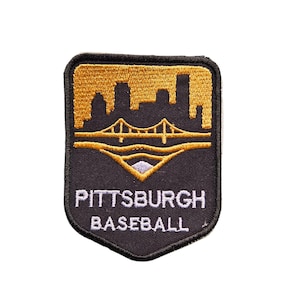 May include: A black and gold embroidered patch featuring the Pittsburgh skyline and a bridge. The patch has the words "PITTSBURGH BASEBALL" in white lettering. The patch is shield-shaped with a black border.