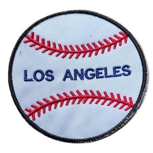 May include: A circular embroidered patch featuring a white baseball design with two curved red stitched seams. The text LOS ANGELES is embroidered in blue capital letters across the center of the patch, which is bordered by a thick black stitched edge.