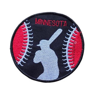 Könnte beinhalten: Ein runder gestickter Aufnäher mit schwarzem Hintergrund und dem Wort MINNESOTA in roter Schrift. Das Design zeigt die hellblaue Silhouette eines Baseballspielers mit Schläger, flankiert von zwei roten Baseballnähten mit hellblauen Stichen.