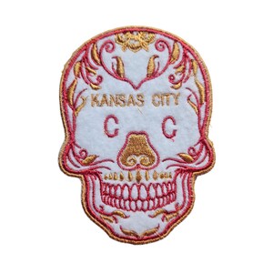 May include: Embroidered patch featuring a white skull with red and gold details. The skull has a floral design and the text "KANSAS CITY C C" is written across the forehead.