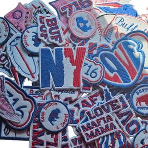 May include: A collection of embroidered patches in various shapes and sizes. The patches feature text like "NY", "Buffalo", and "LOVE MAFIA MAMA", along with other designs in red, white, and blue. Some patches have a vintage aesthetic.