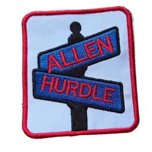 May include: Embroidered patch with a light blue background, featuring a black signpost with two blue signs. The signs read "ALLEN" and "HURDLE" in red letters. The patch has a red border.