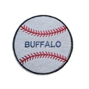 Puede incluir: Parche circular con forma de béisbol, con fondo gris claro y costuras rojas. La palabra "BUFFALO" está bordada en azul. El parche tiene un borde negro.