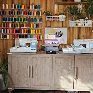 May include: A well-organized sewing room with a wooden cabinet and walls. Two white sewing machines sit on the cabinet, with a variety of colorful thread spools displayed on the wall. Plants and a whiteboard add to the creative space.