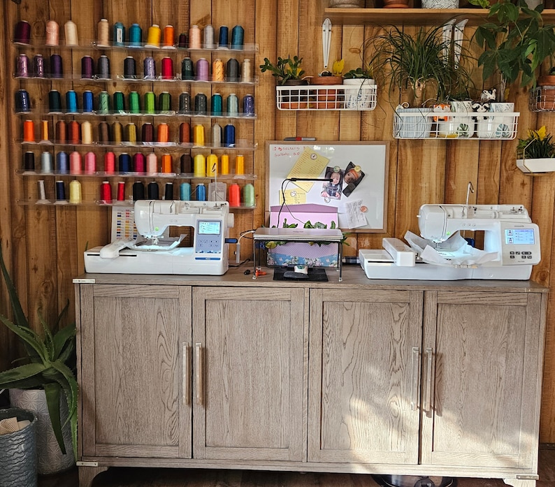 May include: A well-organized sewing room with a wooden cabinet and walls. Two white sewing machines sit on the cabinet, with a variety of colorful thread spools displayed on the wall. Plants and a whiteboard add to the creative space.