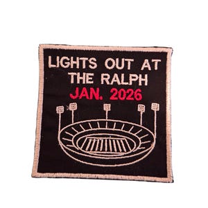 Może przedstawiać: Czarna, haftowana naszywka z białym napisem „LIGHTS OUT AT THE RALPH JAN. 2026”. Naszywka przedstawia projekt stadionu ze światłami. Naszywka ma białą obwódkę.