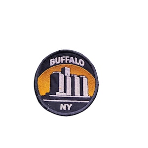 Pode incluir: Um patch bordado redondo com a palavra "BUFFALO" em texto branco arqueado acima de uma ilustração estilizada de edifícios. O patch tem uma borda azul marinho com "NY" em texto branco na parte inferior e um fundo amarelo e preto.