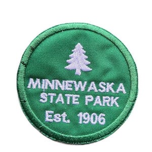 May include: A circular embroidered patch with a green background and a white stitched border. The design features a white pine tree icon above the white text MINNEWASKA STATE PARK and the text Est. 1906 centered at the bottom of the patch.