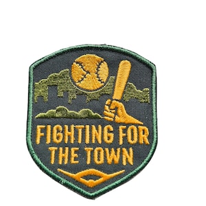 Puede incluir: Parche bordado con temática de béisbol. El parche tiene forma de escudo con un fondo gris oscuro y un borde verde. Presenta una pelota de béisbol, un bate y el texto "FIGHTING FOR THE TOWN" en dorado.