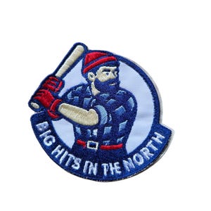 May include: Embroidered patch featuring a lumberjack-style figure in a blue plaid shirt, red hat, and gloves, holding a baseball bat. The patch is circular with the text "BIG HITS IN THE NORTH" in white.