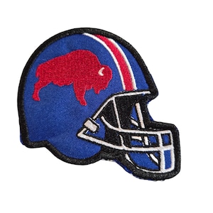 May include: A blue embroidered patch shaped like a football helmet. The helmet features a red buffalo silhouette on the side, a white stripe running over the top, and a black face mask outlined in white thread against a black border.