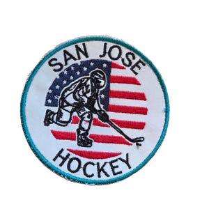 Puede incluir: Un parche redondo con un jugador de hockey en blanco y negro, sobre un fondo de rayas rojas y blancas con estrellas azules. Las palabras "SAN JOSE HOCKEY" están en un diseño circular.