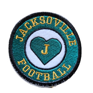 May include: A round, green and gold embroidered patch with the words "JACKSONVILLE FOOTBALL" around a white heart with a gold "J" inside.