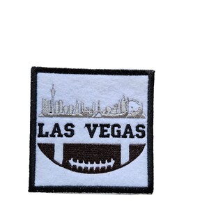 May include: White embroidered patch with a black border. The patch features a silver-colored skyline of Las Vegas with the text "LAS VEGAS" below. A brown football is in the bottom half of the patch.