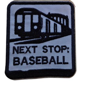 May include: A square embroidered patch with a black border on a light gray background. The design features a black graphic of a subway train car and the text "NEXT STOP: BASEBALL" in bold, black, sans-serif capital letters centered below the train.