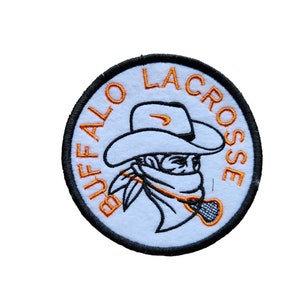 May include: A white embroidered patch with a black outline. The patch features a cowboy wearing a hat and a bandana, holding a lacrosse stick. The text "BUFFALO LACROSSE" is written in orange around the cowboy.