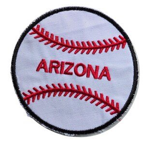 May include: A circular embroidered patch featuring a white baseball design with red stitching details. The word ARIZONA is stitched in red capital letters across the center of the patch, which is finished with a black embroidered border edge.