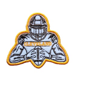 May include: An embroidered patch featuring a football player in a white uniform with a brown helmet and the word "CLEVELAND" on the front. The patch has a yellow outline.