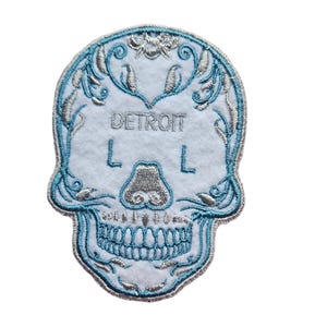 May include: White embroidered patch with a blue sugar skull design. The skull has a floral pattern around the top and the words "DETROIT L L" in the center.