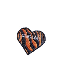 Op de afbeelding: Een zwart en oranje hartvormige patch met het woord "CHICAGO" geborduurd in wit.