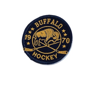 May include: A circular navy blue patch with gold embroidery. The patch features the words "BUFFALO HOCKEY" and the years "1970". A gold buffalo and crossed hockey sticks are in the center. Stars surround the outer edge.