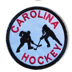 May include: A circular embroidered patch with a white background and a thick black border. The patch features two black silhouettes of hockey players in a face-off position. The red text "CAROLINA" is arched above and "HOCKEY" is arched below.