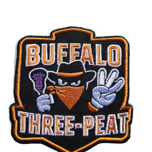Puede incluir: Parche bordado negro y naranja con las palabras "BUFFALO THREE-PEAT". El diseño presenta un personaje de dibujos animados con sombrero y pañuelo, sosteniendo un objeto morado y haciendo el signo de la paz con la mano.