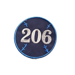 May include: A circular embroidered patch featuring a dark navy blue background with a bright blue stitched border. The number 206 is centered in white thread, surrounded by four small, light blue stitched branch-like symbols positioned at the corners.