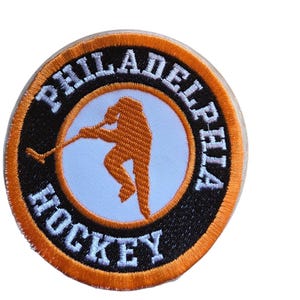 May include: A circular embroidered patch featuring an orange silhouette of a hockey player in the center on a light blue background. The patch has a black border with the white text PHILADELPHIA HOCKEY and an orange outer edge trim.