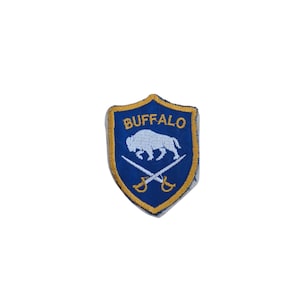 May include: A blue and gold embroidered patch with the word "BUFFALO" at the top. The patch features a white buffalo and crossed swords. The patch is shield-shaped.
