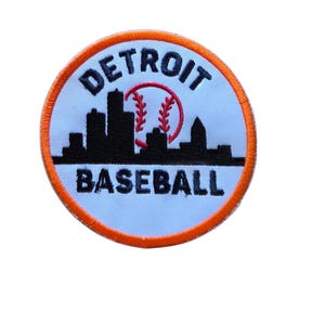 May include: A circular embroidered patch with a white background and an orange border. It features a black silhouette of the Detroit skyline, a red baseball graphic, and the black text DETROIT above and BASEBALL below the skyline design.