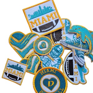 May include: A collection of embroidered patches featuring the Miami Dolphins logo, a heart with the letter D, and the words "Miami Football". The patches are in various colors, including teal, white, and gold.