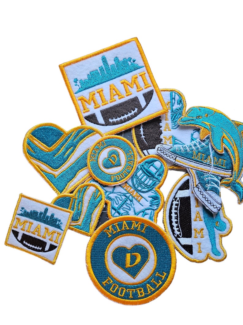 May include: A collection of embroidered patches featuring the Miami Dolphins logo, a heart with the letter D, and the words "Miami Football". The patches are in various colors, including teal, white, and gold.