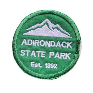 May include: A circular embroidered patch with a green background featuring a light purple mountain graphic at the top. The text reads ADIRONDACK STATE PARK in white capital letters, with the text Est. 1892 printed in white below the main title.