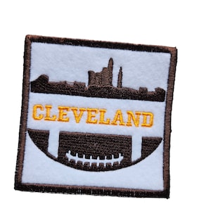 May include: Brown and white embroidered patch featuring a brown silhouette of the Cleveland skyline and a brown football with yellow laces. The text "CLEVELAND" is embroidered in yellow.