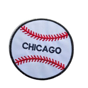 May include: A circular embroidered patch featuring a white baseball design with red stitching details. The word CHICAGO is stitched in dark blue capital letters across the center of the patch, which is finished with a black stitched border.