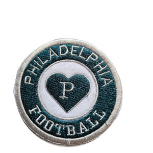 May include: A round, white and teal embroidered patch with the words "Philadelphia Football" in a circle around a white heart with a teal "P" inside.