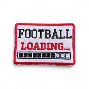 May include: A rectangular embroidered patch with a white background and a red border. The text FOOTBALL is in black, and LOADING... is in red. Below the text is a loading bar graphic with black and white segments, indicating partial progress.