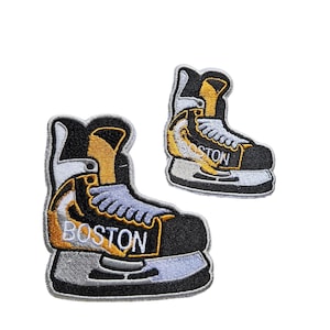 Könnte beinhalten: Zwei gestickte Bügelbilder von Schlittschuhen mit dem Wort "BOSTON" darauf. Das größere Bügelbild ist schwarz, weiß und gold. Das kleinere Bügelbild ist schwarz, weiß und gelb.