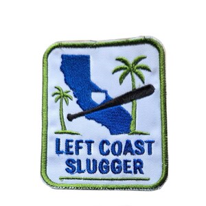 May include: A rectangular embroidered patch with a white background and a green border. It features a blue silhouette of California, two green palm trees, and a black baseball bat. The text "LEFT COAST SLUGGER" is embroidered in blue letters.