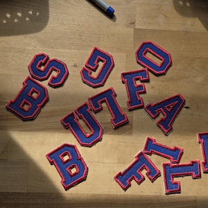 May include: A collection of blue and red embroidered letter patches spelling "BUFFALO" are arranged on a wooden surface. The patches feature a blue base with red edging, in a classic varsity style.