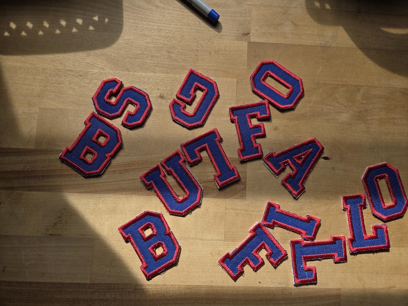 May include: A collection of blue and red embroidered letter patches spelling "BUFFALO" are arranged on a wooden surface. The patches feature a blue base with red edging, in a classic varsity style.