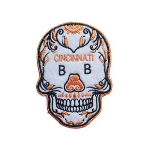 Puede incluir: Parche blanco bordado con un diseño de calavera negra y naranja. La calavera tiene un patrón floral y el texto "CINCINNATI B B" está escrito en la frente.
