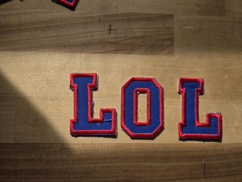 May include: Embroidered patches spelling out "LOL" in blue with red trim. The patches are arranged on a wooden surface. The letters have a collegiate style.
