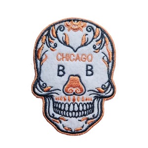 May include: An embroidered patch featuring a white skull with orange floral details. The skull has a black outline and black teeth. The text "CHICAGO B B" is written in black on the skull's forehead.