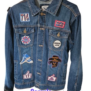 May include: A blue denim jacket with various embroidered patches. Patches include the number "716", the words "MAFIA MAMA", a thumbs-up with "17", and "Buffalo NLL Champs 23 24 Back to Back". The jacket is hanging on a black hanger. The text "Sample only" is also visible.