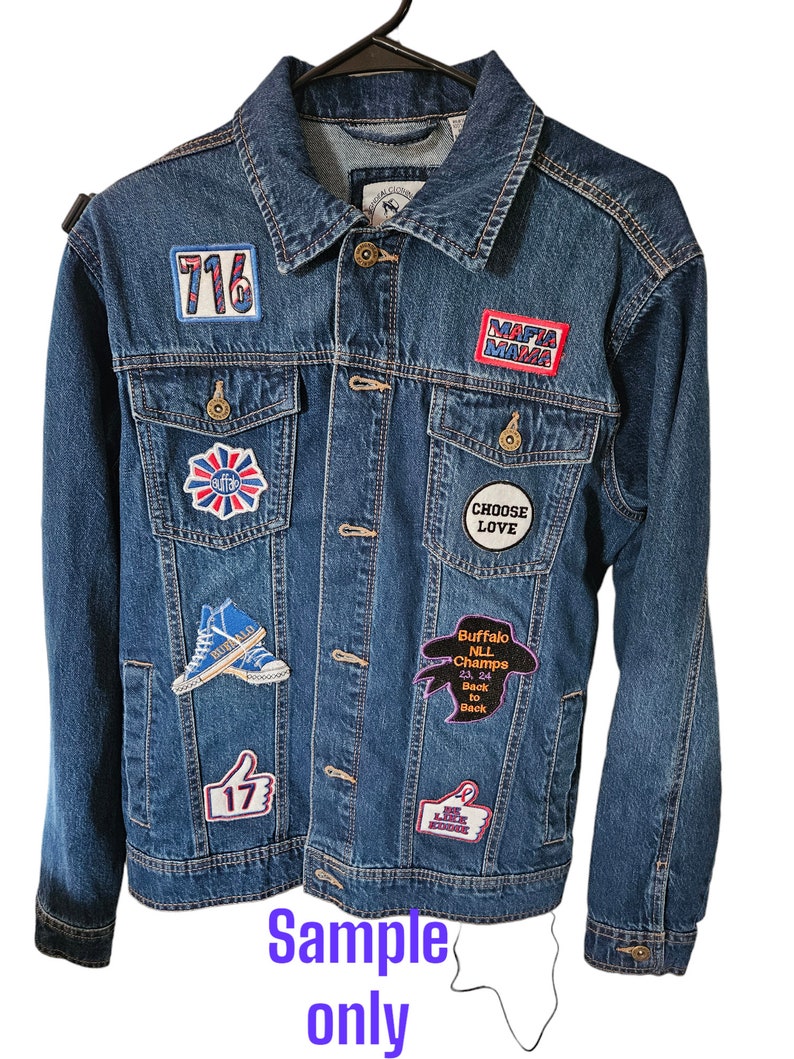 May include: A blue denim jacket with various embroidered patches. Patches include the number "716", the words "MAFIA MAMA", a thumbs-up with "17", and "Buffalo NLL Champs 23 24 Back to Back". The jacket is hanging on a black hanger. The text "Sample only" is also visible.