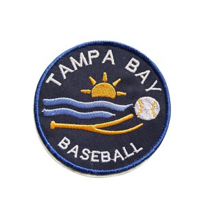 May include: A navy blue circular patch with white embroidered text that reads "TAMPA BAY BASEBALL". The patch features a yellow sun, blue waves, a baseball, and a baseball bat.