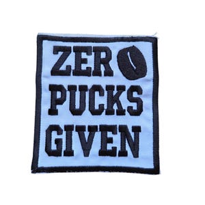 May include: A light blue patch with black embroidered text that reads "ZERO PUCKS GIVEN". The word "pucks" has a hockey puck graphic in place of the letter "O". The patch has a black border.